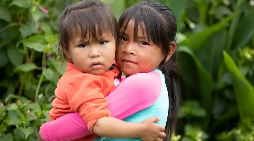 Older girl gently holding her younger sister in her arms outdoors in greenery